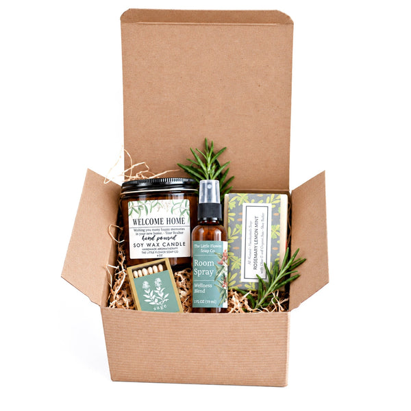 A gift box set containing a soy wax candle, a room spray, a matchbox, and a bath bomb, arranged in a brown kraft box with shredded paper on a white background.
